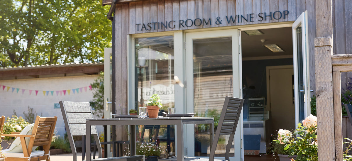 A rustic wooden building with a sign reading 'TASTING ROOM & WINE SHOP' has patio seating outside, including metal tables and chairs, with trees and a garden visible in the background.