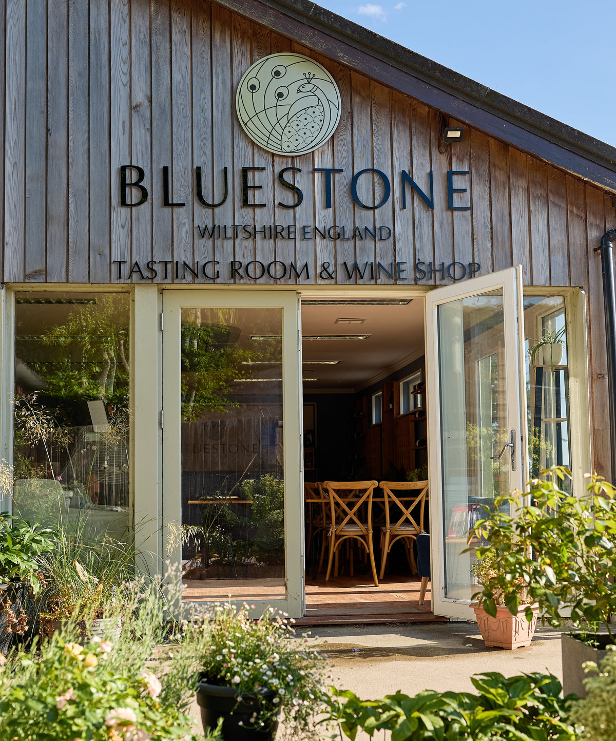 Bluestone Wilshire England Tasting Room & Wine Shop exterior, featuring a rustic wooden facade, large glass doors, and potted green plants in the foreground under a blue sky.